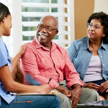 A caregiver and a daughter chatting warmly with an older woman resting on the sofa at home